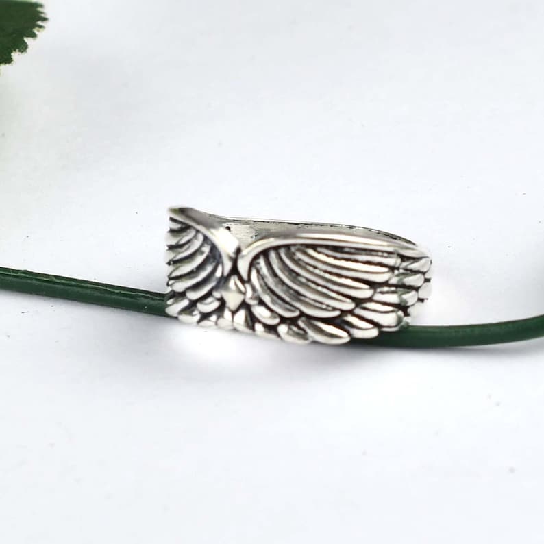May include: A silver-toned ring featuring a detailed, symmetrical wing design that wraps around the band. The metal has a polished finish with darkened recessed areas to highlight the individual feather textures carved into the surface.