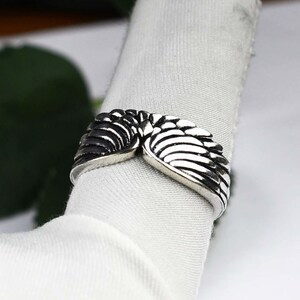 May include: A silver-toned metal ring featuring a detailed design of two symmetrical wings that meet in the center. The surface of the wings has deep, dark oxidized grooves to highlight the individual feather textures across the band.