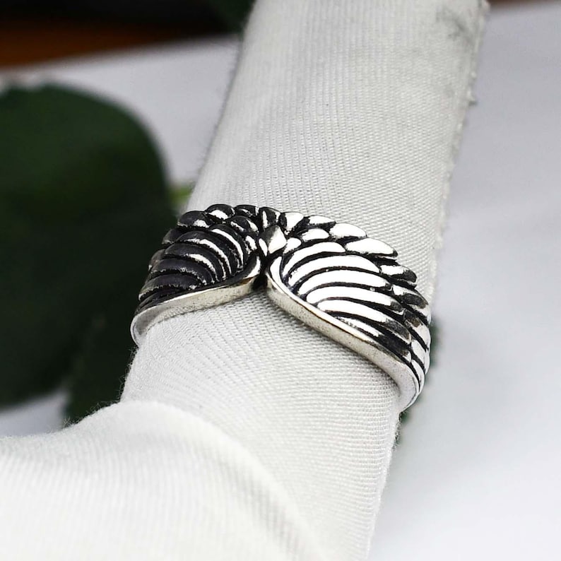 May include: A silver-toned metal ring featuring a detailed design of two symmetrical wings that meet in the center. The surface of the wings has deep, dark oxidized grooves to highlight the individual feather textures across the band.