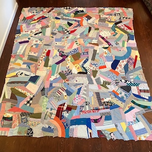 May include: A large, colorful quilt composed of various fabric scraps in a patchwork design. The quilt features a mix of patterns, including floral, plaid, and solid colors, creating a vibrant and textured appearance. The quilt is displayed on a wooden floor.