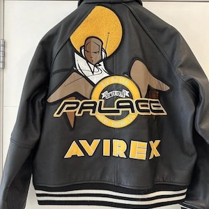 May include: Black leather jacket with a large graphic on the back featuring a seated figure with an afro, and the words "AVIREX PALACE NEW YORK NY" in yellow and white. The jacket has a black and white striped hem.