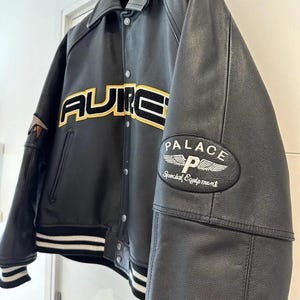 May include: Black leather jacket with the word "AURIC" in yellow and black lettering across the chest. The jacket has a white and black striped hem and cuffs, and a "PALACE Special Equipment" patch on the sleeve.