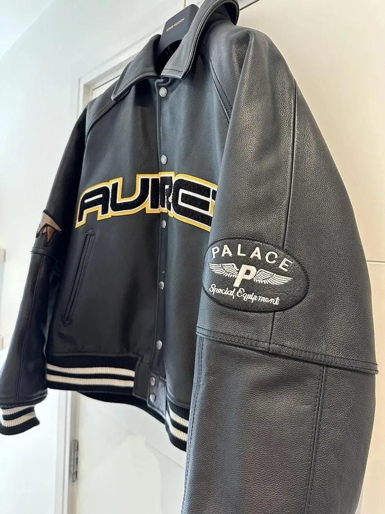 May include: Black leather jacket with the word "AURIC" in yellow and black lettering across the chest. The jacket has a white and black striped hem and cuffs, and a "PALACE Special Equipment" patch on the sleeve.