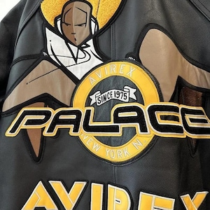 May include: Black leather jacket with yellow and white embroidered patches. Features the text "PALACE NEW YORK N" and "AVIREX SINCE 1975". A graphic of a person with a yellow halo is also visible.