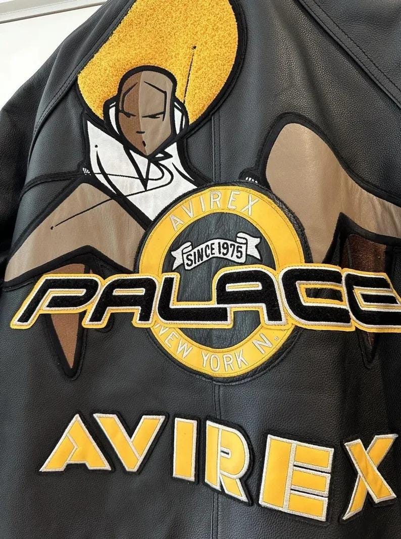 May include: Black leather jacket with yellow and white embroidered patches. Features the text "PALACE NEW YORK N" and "AVIREX SINCE 1975". A graphic of a person with a yellow halo is also visible.