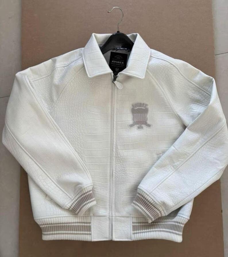Op de afbeelding: Een witte leren jas met een getextureerd, krokodilachtig patroon. De jas heeft een ritssluiting, een kraag en een logopatch op de borst. Geribde manchetten en zoom hebben beige en witte strepen. Hangt aan een zwarte hanger.