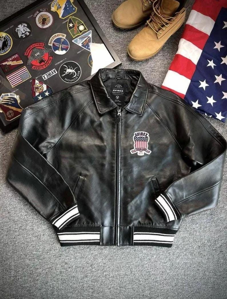 Pu&ograve; includere: Giacca in pelle nera con zip, colletto e toppa sul petto. La giacca ha strisce bianche sui polsini e sull'orlo. L'immagine mostra anche stivali marroni e una bandiera americana.