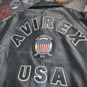 Pu&ograve; includere: Giacca in pelle nera con scritte bianche "AVIREX" e "USA". Una toppa circolare presenta le parole "ALL AMERICANS" e "ESTABLISHED 1975" con un disegno della bandiera americana. La giacca ha un classico stile bomber.