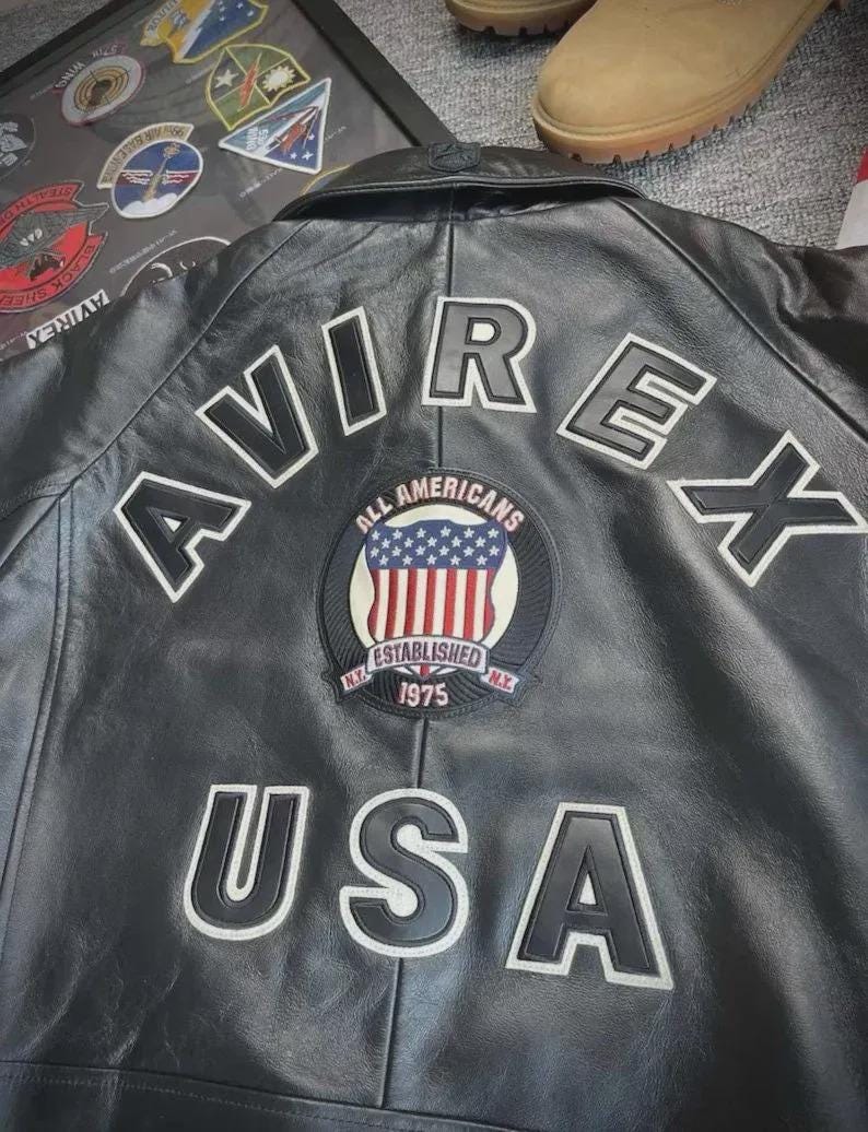 Pu&ograve; includere: Giacca in pelle nera con scritte bianche "AVIREX" e "USA". Una toppa circolare presenta le parole "ALL AMERICANS" e "ESTABLISHED 1975" con un disegno della bandiera americana. La giacca ha un classico stile bomber.