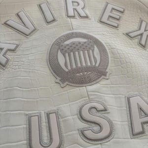 Op de afbeelding: Cr&egrave;me leren jas met een krokodillenleer effect. De jas heeft de letters "VIREX" en "USA" in reli&euml;f. Een cirkelvormige patch met een Amerikaans vlagontwerp is ook zichtbaar.