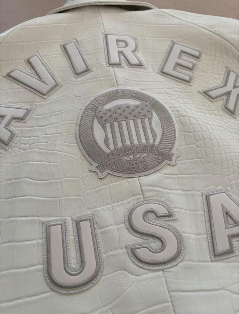 Op de afbeelding: Cr&egrave;me leren jas met een krokodillenleer effect. De jas heeft de letters "VIREX" en "USA" in reli&euml;f. Een cirkelvormige patch met een Amerikaans vlagontwerp is ook zichtbaar.
