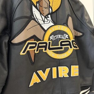 May include: Black leather jacket with a large graphic on the back. The graphic features a stylized figure with a yellow afro and the words "PALACE NEW YORK" and "AVIREX SINCE 1975" in yellow and white lettering. The jacket has a striped hem.