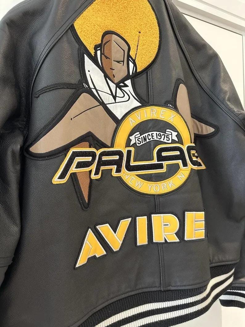 May include: Black leather jacket with a large graphic on the back. The graphic features a stylized figure with a yellow afro and the words "PALACE NEW YORK" and "AVIREX SINCE 1975" in yellow and white lettering. The jacket has a striped hem.