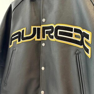 May include: Black leather jacket with the word "AUIRCE" in large, yellow-outlined, black-filled letters across the chest. The jacket has a button-up front, a collar, and two front pockets. The jacket has a classic varsity style.
