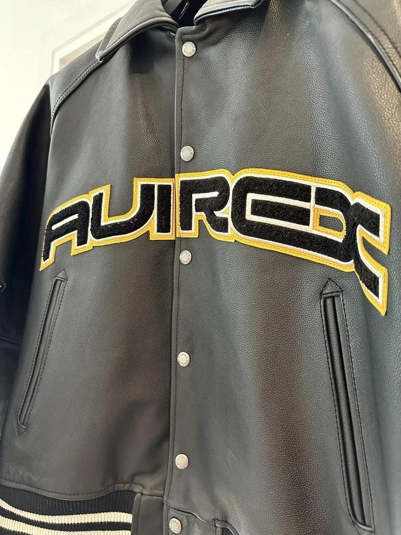 May include: Black leather jacket with the word "AUIRCE" in large, yellow-outlined, black-filled letters across the chest. The jacket has a button-up front, a collar, and two front pockets. The jacket has a classic varsity style.