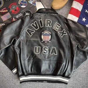 Pu&ograve; includere: Giacca in pelle nera con la scritta "AVIREX" in lettere bianche sul retro. Una toppa circolare con la bandiera americana e le parole "ALL AMERICANS ESTABLISHED 1975" &egrave; sotto. La giacca ha un orlo a righe bianche e nere.