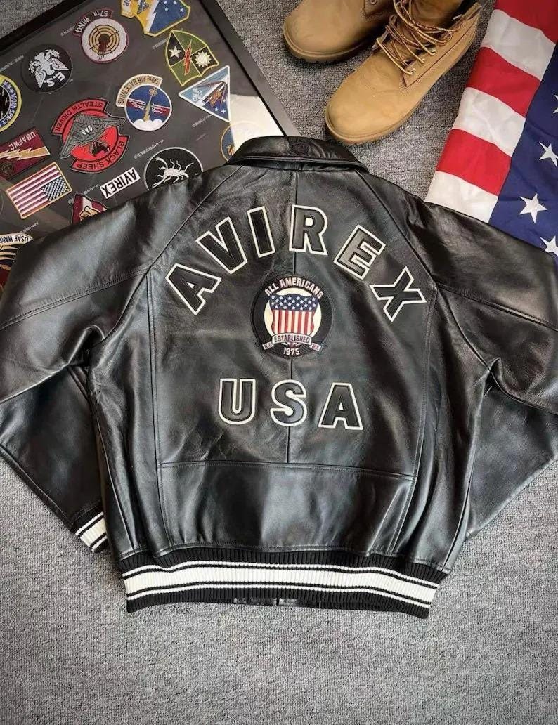 Pu&ograve; includere: Giacca in pelle nera con la scritta "AVIREX" in lettere bianche sul retro. Una toppa circolare con la bandiera americana e le parole "ALL AMERICANS ESTABLISHED 1975" &egrave; sotto. La giacca ha un orlo a righe bianche e nere.