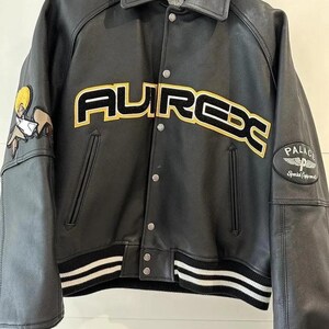 May include: Black leather jacket with the word "AUREX" in yellow and black lettering across the chest. The jacket has a collar, snap closures, and white and black striped cuffs and hem. Patches are on the sleeves.