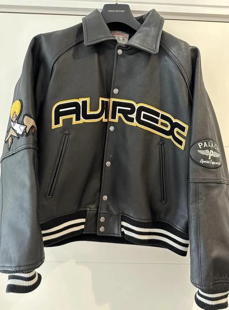 May include: Black leather jacket with the word "AUREX" in yellow and black lettering across the chest. The jacket has a collar, snap closures, and white and black striped cuffs and hem. Patches are on the sleeves.