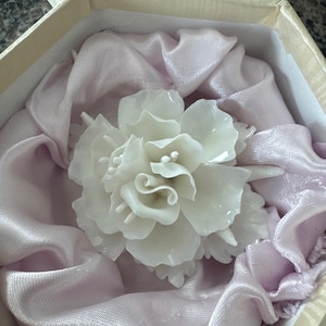 May include: A white, sculpted flower, possibly a brooch or hair accessory, rests on a bed of pale pink satin fabric inside a hexagonal, cream-colored box. The flower has detailed petals and is the focal point.