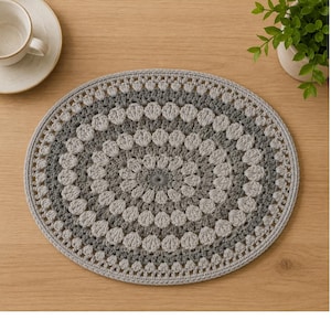 May include: A round, crocheted placemat featuring concentric circular patterns in alternating shades of light gray, medium gray, and dark charcoal gray. The design consists of repeating textured stitch motifs radiating from the center.
