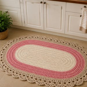May include: An oval-shaped crochet rug featuring a central cream-colored section, a surrounding band of pink, and an outer border of beige with a decorative scalloped edge. The rug is constructed with visible yarn stitches in a textured pattern.