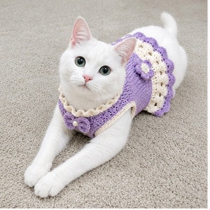 May include: A white cat wearing a handmade lavender and cream-coloured knitted dress. The dress features a scalloped skirt, a bow detail, and a flower accent. The cat is lying down on a light grey carpet.