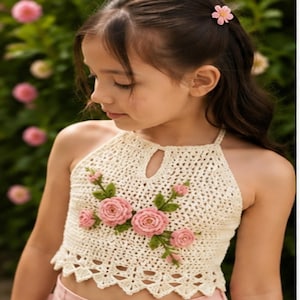 Puede incluir: Un top halter de ganchillo color crema con escote tipo cerradura y un grupo decorativo de rosas de ganchillo rosas con hojas verdes en la parte delantera. El dobladillo tiene un diseño festoneado y la tela un patrón calado.
