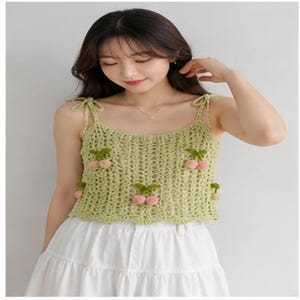 May include: A light green crochet top with cherry embellishments. The top features a scoop neckline and tie straps. The cherries are pink with green leaves. The top is paired with a white skirt.