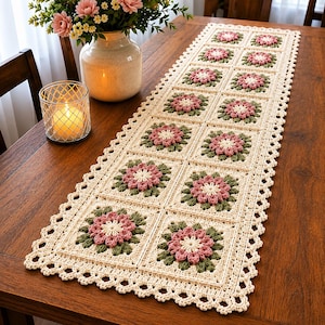 Può includere: Runner da tavolo all'uncinetto color crema con un motivo floreale ripetuto. Ogni quadrato presenta un motivo floreale rosa e verde. Il runner ha un bordo smerlato ed è esposto su un tavolo di legno. Un vaso di fiori e una candela sono anche sul tavolo.