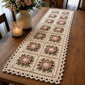 Pode incluir: Caminho de mesa de crochê creme com um padrão floral. O caminho de mesa apresenta motivos quadrados com desenhos de flores rosa e verde, bordados por uma borda ondulada. O caminho de mesa é exibido em uma mesa de madeira.