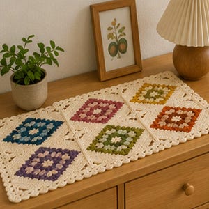 May include: A cream-colored, rectangular crochet table runner with six colorful diamond-shaped patterns. The diamonds feature shades of blue, purple, green, yellow, and orange. The runner is placed on a wooden surface, with a small potted plant and a framed picture in the background.