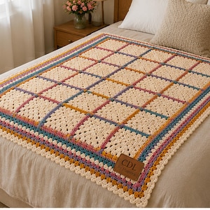 Könnte beinhalten: Eine cremefarbene Häkeldecke mit einem Gitter aus quadratischen Motiven, die durch blaue, rosa und gelbe Garnränder getrennt sind. Die Decke hat einen gewellten Rand und einen braunen rechteckigen Lederaufnäher mit dem Text CDL by Himera.