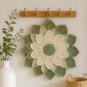 May include: A circular crochet wall hanging featuring a layered floral design with a sage green center, cream-colored inner petals, and outer sage green leaves. The piece is suspended from a wooden hook rack by a small loop at the top center.