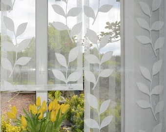 May include: White sheer window curtains with a leaf pattern. The curtains are hanging in front of a window with a view of green foliage and a vase of yellow tulips. The leaf design is repeated across the panels.