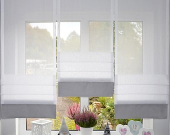 May include: Three white sheer roman shades with solid gray bottom panels hanging in a window. The shades feature horizontal folds and are positioned above a windowsill containing a white lantern, two gnome figurines, and three heart-shaped signs.