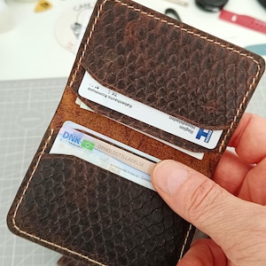 Puede incluir: Un tarjetero de cuero marrón oscuro con un patrón texturizado similar a la piel de serpiente y costuras claras. La cartera cuenta con dos ranuras visibles que contienen tarjetas de identificación, incluyendo una marcada Københavns Kommune y otra OPHOLDSTILLADELSE.