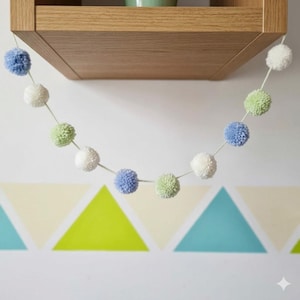 Könnte beinhalten: Eine Girlande mit weißen, hellblauen und grünen Pompons, die von einem Holzregal hängt. Die flauschigen Pompons sind an einer Schnur befestigt und bilden ein dekoratives Element. Der Hintergrund zeigt ein geometrisches Dreieckmuster.