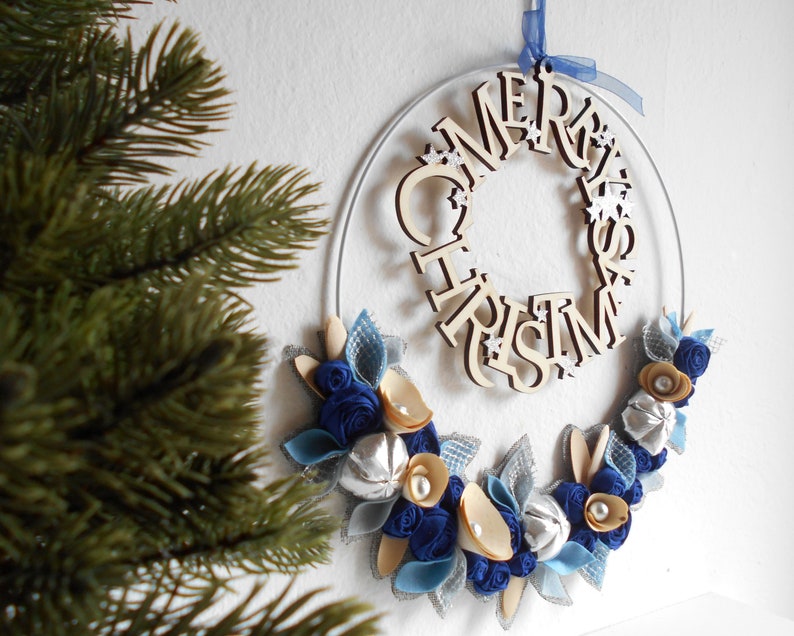 white metal hoop decorated with a floral arrangement composed of blue fabric roses, felt leaves, wooden flowers, and silver details.
A wooden laser-cut MERRY CHRISTMAS SIGN takes place on the top.