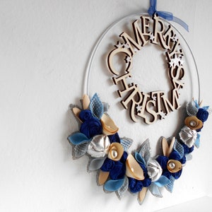 white metal hoop decorated with a floral arrangement composed of blue fabric roses, felt leaves, wooden flowers, and silver details.
A wooden laser-cut MERRY CHRISTMAS SIGN takes place on the top.