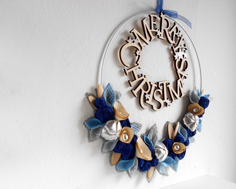 white metal hoop decorated with a floral arrangement composed of blue fabric roses, felt leaves, wooden flowers, and silver details.
A wooden laser-cut MERRY CHRISTMAS SIGN takes place on the top.