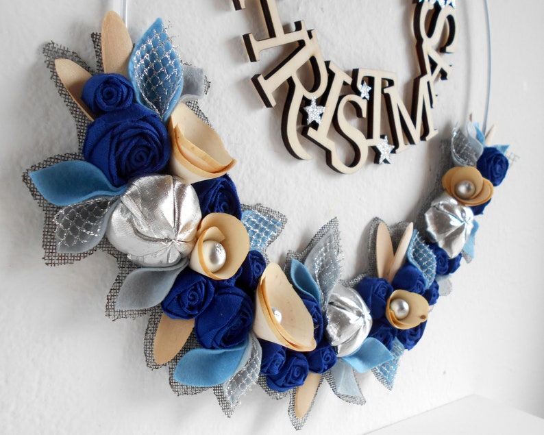 white metal hoop decorated with a floral arrangement composed of blue fabric roses, felt leaves, wooden flowers, and silver details.
A wooden laser-cut MERRY CHRISTMAS SIGN takes place on the top.