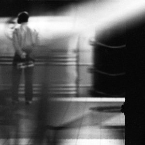 Puede incluir: Fotografía en blanco y negro de una persona en un espacio borroso de aspecto industrial. La persona se ve de espaldas, con las manos entrelazadas. Una fuerte fuente de luz crea sombras y reflejos, acentuando el ambiente.