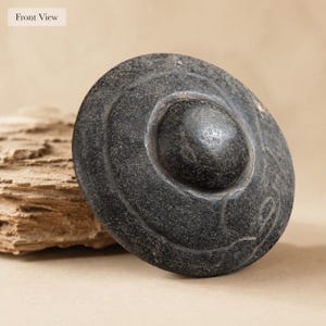 May include: A circular dark gray stone object with a smooth, raised central dome and concentric circular grooves etched into the surface. The stone has a textured, weathered appearance with visible natural variations in the dark gray material.