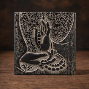 Può includere: Una placca quadrata in pietra grigio scuro con un rilievo scolpito di due mani in un gesto mudra. Le mani sono adornate con una fila di grani di preghiera che si adagia sui palmi e sulle dita, su uno sfondo strutturato.