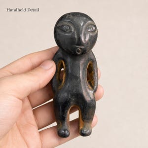 May include: A small, dark, smooth-surfaced figurine with a rounded head, almond-shaped eyes, and a small circular mouth. The figure has an abstract, stylized human form with open spaces between the arms and torso, featuring a dark exterior finish.