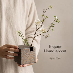 May include: A square, dark gray ceramic vase with a textured, speckled surface and a single opening at the top. A thin, brown branch with small green leaves and two white flowers is placed inside. The text Elegant Home Accent Square Vase is visible.