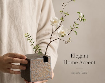 May include: A square, dark gray ceramic vase with a textured, speckled surface and a single opening at the top. A thin, brown branch with small green leaves and two white flowers is placed inside. The text Elegant Home Accent Square Vase is visible.