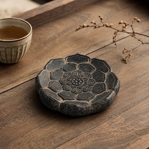 May include: A dark gray stone coaster shaped like a blooming lotus flower with intricate carved petal details on the surface. The coaster has a textured, natural finish and a circular form, providing a stable base for small items or tea cups.