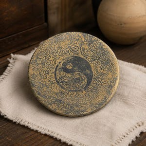 May include: A circular, flat, metallic-toned object featuring a central yin-yang symbol surrounded by an intricate, repeating floral pattern in dark gray and muted gold. The surface has a weathered, textured appearance with visible signs of aging.